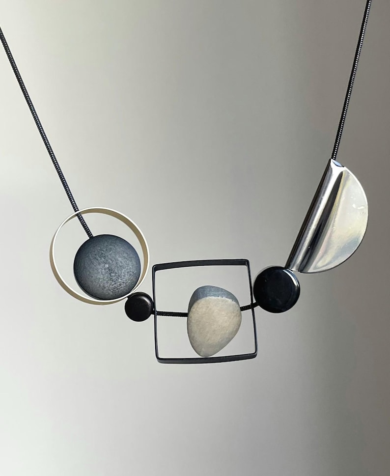May include: A necklace featuring a black cord strung with geometric elements including a silver circle containing a dark gray sphere, a black square frame holding a light gray stone, two small black beads, and a polished silver crescent shape.