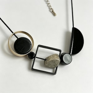 May include: A necklace featuring a black cord strung with geometric elements including a gold-toned circle enclosing a black disc, a black square frame containing a textured grey stone, a small black bead, and a black semi-circular piece.