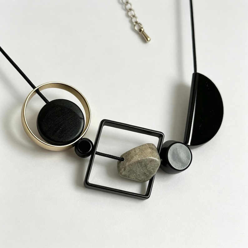 May include: A necklace featuring a black cord strung with geometric elements including a gold-toned circle enclosing a black disc, a black square frame containing a textured grey stone, a small black bead, and a black semi-circular piece.