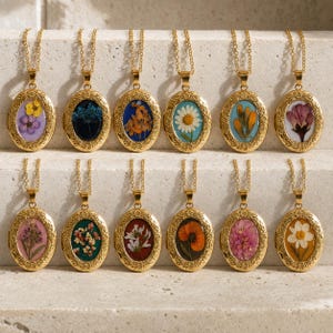 May include: A collection of twelve oval gold-tone pendants on matching chains, each featuring a unique pressed flower encased in resin. The frames have an ornate, textured border, and the floral specimens include daisies, poppies, and various wildflowers.