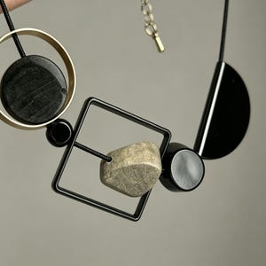 May include: A necklace featuring a black cord strung with geometric elements including a gold-toned circle enclosing a black disc, a black square frame containing a textured beige stone, and a black semi-circle, finished with a metal chain extender.
