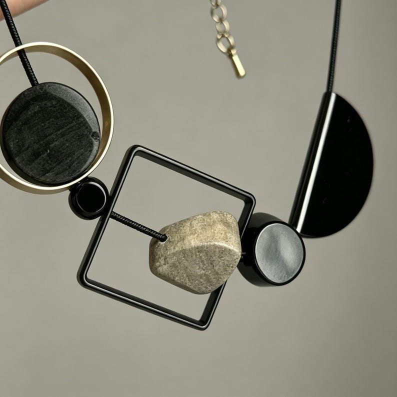 May include: A necklace featuring a black cord strung with geometric elements including a gold-toned circle enclosing a black disc, a black square frame containing a textured beige stone, and a black semi-circle, finished with a metal chain extender.