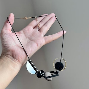 May include: A necklace featuring a thin black cord with a gold-toned metal clasp. The pendant consists of a black semi-circle, a small black cylinder, a smooth beige stone, a black square frame, and a gold-toned circle with a black disc inside.