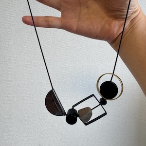 May include: A necklace featuring a black cord and a series of geometric pendants, including a black semi-circle, a small black cylinder, a black square frame containing a textured tan stone, and a gold-toned circle with a black circular center.
