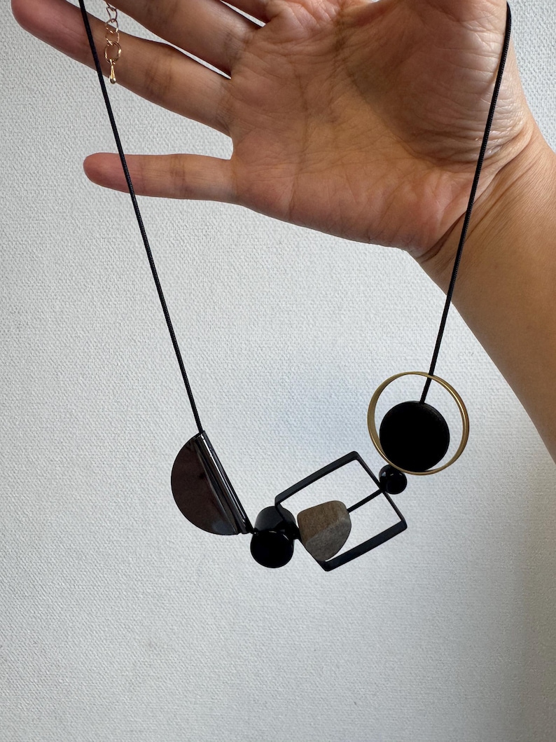 May include: A necklace featuring a black cord and a series of geometric pendants, including a black semi-circle, a small black cylinder, a black square frame containing a textured tan stone, and a gold-toned circle with a black circular center.