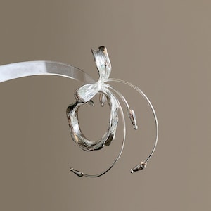 May include: A silver-toned metallic ear cuff featuring a textured, organic circular shape that wraps around the ear. The design includes a prominent, elongated petal-like structure extending upward and three thin, curved wires ending in small beads.