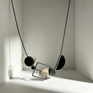 May include: A necklace featuring a black cord strung with geometric elements including a gold-toned circle, a black square frame, a black semi-circle, and small black cylinders, accented by a single textured, light-colored stone bead.