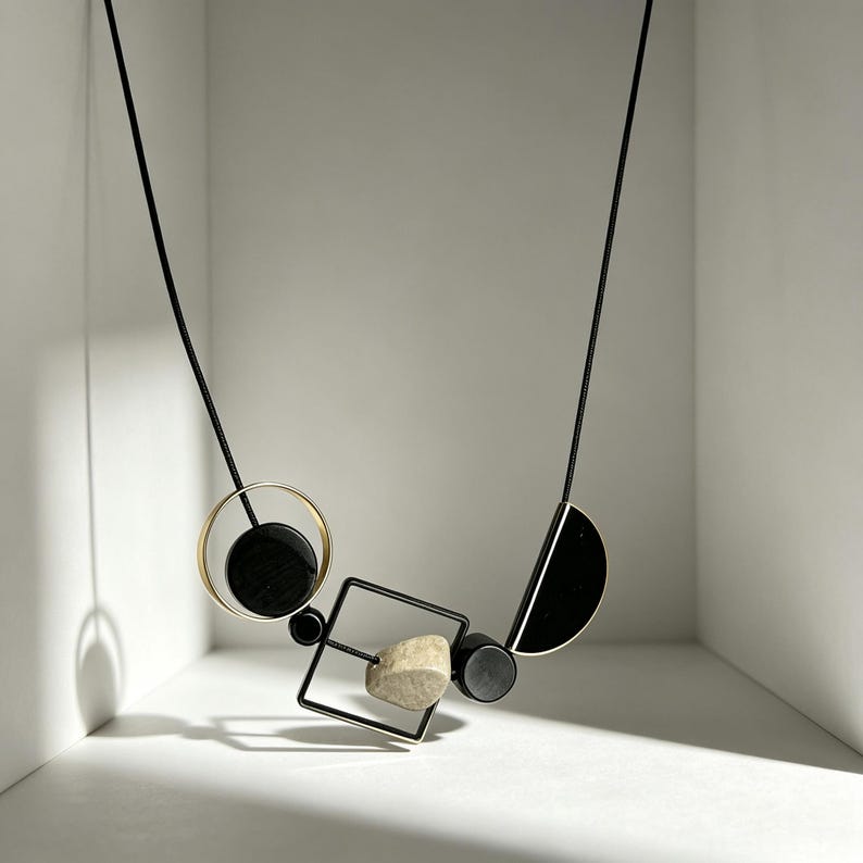 May include: A necklace featuring a black cord strung with geometric elements including a gold-toned circle, a black square frame, a black semi-circle, and small black cylinders, accented by a single textured, light-colored stone bead.