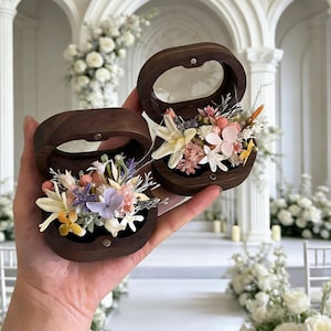 Könnte beinhalten: Zwei offene, dunkle Holz-Ringboxen, jede gefüllt mit einem bunten Sortiment an Trockenblumen. Die Boxen werden in einer Hand gehalten, mit einer Hochzeitszeremonie im Hintergrund. Die Blumen umfassen rosa, weiße, gelbe und lila Blüten.