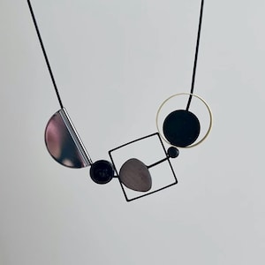 May include: A necklace featuring a thin black cord suspending a geometric arrangement of a metallic silver semicircle, a small black bead, a black square frame containing a gray pebble-like shape, and a black circle inside a thin gold ring.