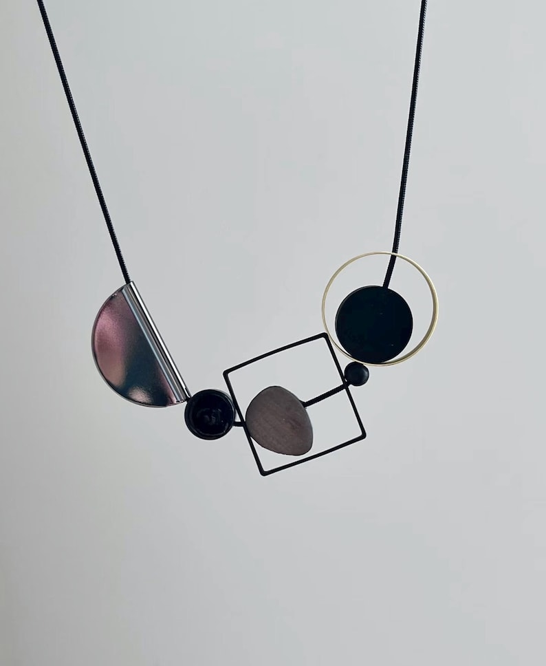 May include: A necklace featuring a thin black cord suspending a geometric arrangement of a metallic silver semicircle, a small black bead, a black square frame containing a gray pebble-like shape, and a black circle inside a thin gold ring.