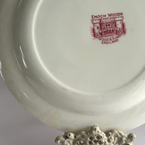 Op de afbeelding: Cr&egrave;mewitte keramische plaat met een rode stempel met de tekst "ENOCH WOODS CASTLES STOKESAY WOOD & SONS ENGLAND". De plaat wordt getoond op een decoratieve, cr&egrave;mewitte metalen standaard.