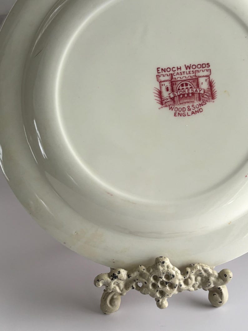 Op de afbeelding: Cr&egrave;mewitte keramische plaat met een rode stempel met de tekst "ENOCH WOODS CASTLES STOKESAY WOOD & SONS ENGLAND". De plaat wordt getoond op een decoratieve, cr&egrave;mewitte metalen standaard.