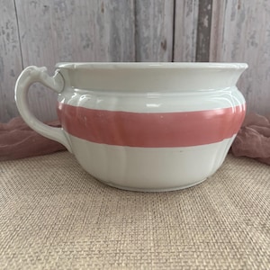 May include: White ceramic bowl with a wide, horizontal coral-colored stripe. The bowl has a handle and a slightly flared rim. The background is a light, neutral color, suggesting a vintage or farmhouse aesthetic.