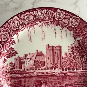Op de afbeelding: Decoratieve plaat met een rood-witte kasteelsc&egrave;ne. De plaat heeft een bloemenrand met rozen en een gedetailleerd landschap met een kasteel, bomen en water. De plaat is waarschijnlijk gemaakt van keramiek.