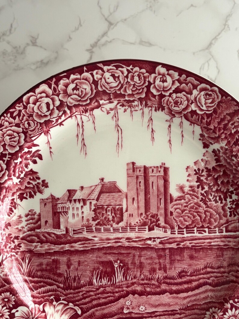 Op de afbeelding: Decoratieve plaat met een rood-witte kasteelsc&egrave;ne. De plaat heeft een bloemenrand met rozen en een gedetailleerd landschap met een kasteel, bomen en water. De plaat is waarschijnlijk gemaakt van keramiek.
