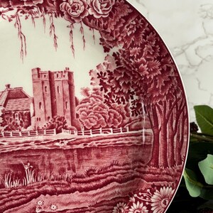 Op de afbeelding: Decoratieve bord met een bordeauxrood en wit landschapsontwerp. Het bord toont een kasteel, bomen en een rivierlandschap, omgeven door bloemen- en rozenpatronen. Het bord heeft een gedetailleerde, vintage esthetiek.