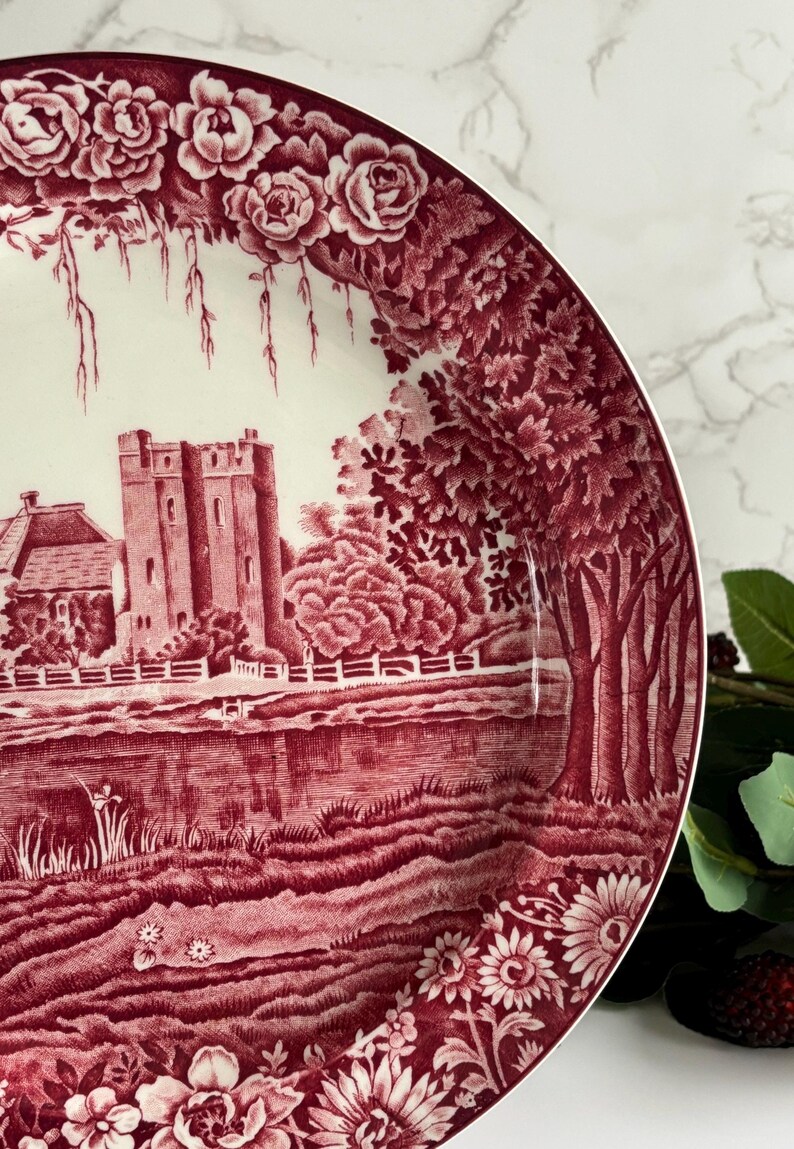 Op de afbeelding: Decoratieve bord met een bordeauxrood en wit landschapsontwerp. Het bord toont een kasteel, bomen en een rivierlandschap, omgeven door bloemen- en rozenpatronen. Het bord heeft een gedetailleerde, vintage esthetiek.