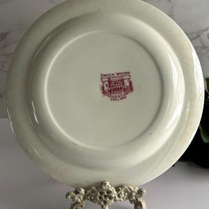 Op de afbeelding: Een witte keramische plaat met een opstaande rand, met een rood-wit wapen met de tekst "ENOCH WOODS" en "WOOD & SONS ENGLAND". De plaat wordt gepresenteerd op een decoratieve standaard.