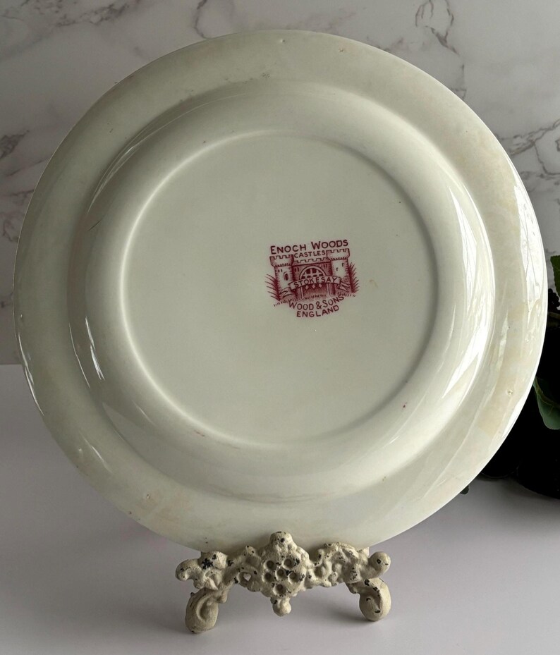 Op de afbeelding: Een witte keramische plaat met een opstaande rand, met een rood-wit wapen met de tekst "ENOCH WOODS" en "WOOD & SONS ENGLAND". De plaat wordt gepresenteerd op een decoratieve standaard.