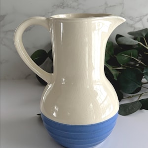 May include: Cream ceramic jug with a blue, ribbed base and a curved handle. The jug has a spout for pouring. The background is a marble-effect surface with green foliage.