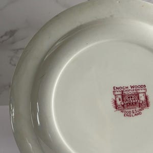 Op de afbeelding: Cr&egrave;mekleurige keramische plaat met een brede rand. De plaat heeft een rood-wit embleem met de tekst "ENOCH WOODS CASTLE STOKESAY WOOD & SONS ENGLAND". De plaat heeft een glanzende afwerking en is waarschijnlijk vintage.