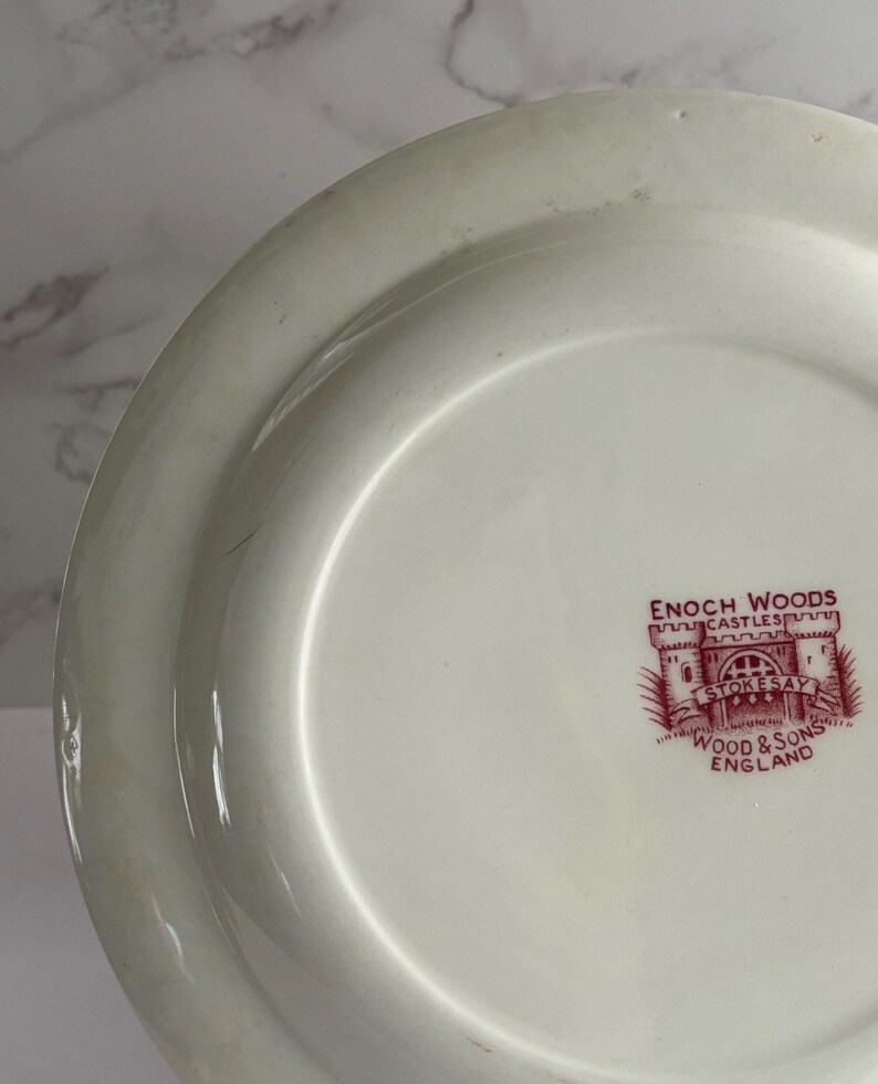 Op de afbeelding: Cr&egrave;mekleurige keramische plaat met een brede rand. De plaat heeft een rood-wit embleem met de tekst "ENOCH WOODS CASTLE STOKESAY WOOD & SONS ENGLAND". De plaat heeft een glanzende afwerking en is waarschijnlijk vintage.