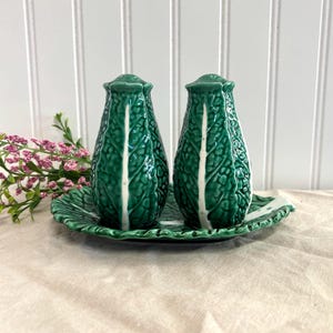 May include: A ceramic salt and pepper shaker set. The shakers are shaped like green cabbage heads with a white stripe down the center. They sit on a matching green leaf-shaped tray. The set is on a beige surface with a white background.