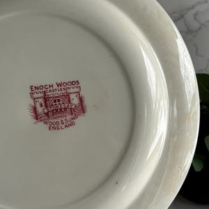 Op de afbeelding: Cr&egrave;mekleurige keramische plaat met een rood embleem met de tekst "ENOCH WOODS CASTLES STOKESAY WOOD & SONS ENGLAND". De plaat heeft een licht verhoogde rand en een glad oppervlak, met een vintage uitstraling.