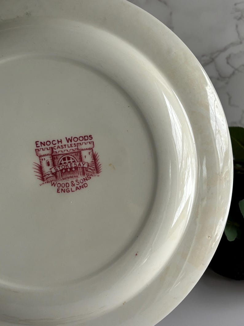 Op de afbeelding: Cr&egrave;mekleurige keramische plaat met een rood embleem met de tekst "ENOCH WOODS CASTLES STOKESAY WOOD & SONS ENGLAND". De plaat heeft een licht verhoogde rand en een glad oppervlak, met een vintage uitstraling.