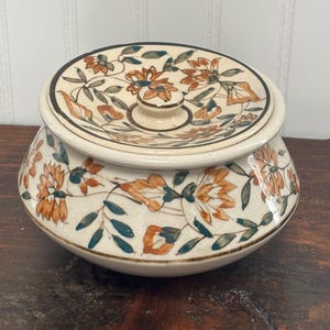 May include: A cream-colored ceramic jar with a lid, adorned with orange and green floral patterns. The jar has a rounded shape with a lid that fits snugly on top. The floral design is repeated on the lid and the body of the jar.