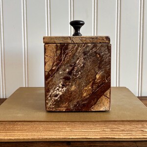 May include: A square box made of brown and tan marbled stone with a smooth, polished finish. The box features a removable lid topped with a small, dark, rounded knob handle. The stone displays natural veining patterns in shades of deep chocolate brown, cream, and tan throughout the exterior surfaces.