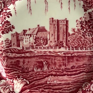 Op de afbeelding: Een decoratieve rode en witte plaat met een kasteelsc&egrave;ne. De plaat heeft een gedetailleerde illustratie van een kasteel, bomen en water, omlijst door een bloemenrand. Het ontwerp is in een klassieke, vintage stijl.