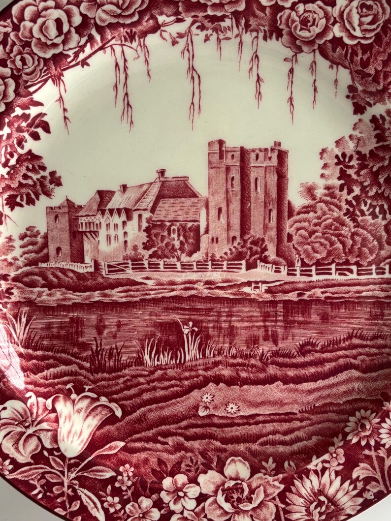 Op de afbeelding: Een decoratieve rode en witte plaat met een kasteelsc&egrave;ne. De plaat heeft een gedetailleerde illustratie van een kasteel, bomen en water, omlijst door een bloemenrand. Het ontwerp is in een klassieke, vintage stijl.