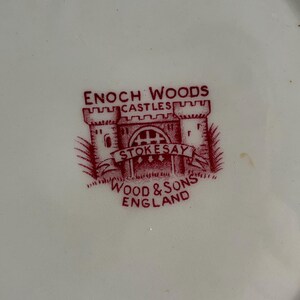 Op de afbeelding: Close-up van een wit bord met een rood Enoch Woods logo. Het logo toont een kasteel illustratie met de woorden "CASTLES" en "STOKESAY" boven en onder het kasteel. De woorden "WOOD & SONS ENGLAND" zijn ook zichtbaar.