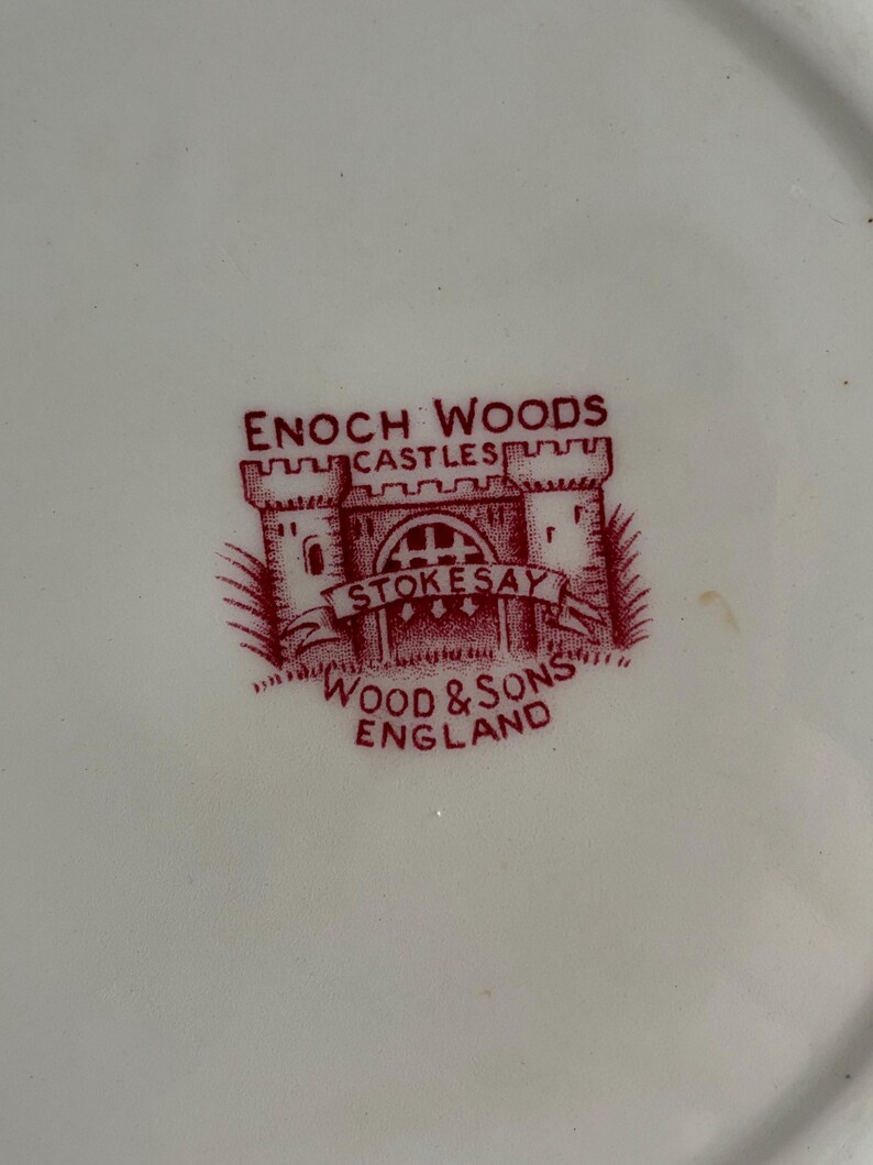 Op de afbeelding: Close-up van een wit bord met een rood Enoch Woods logo. Het logo toont een kasteel illustratie met de woorden "CASTLES" en "STOKESAY" boven en onder het kasteel. De woorden "WOOD & SONS ENGLAND" zijn ook zichtbaar.