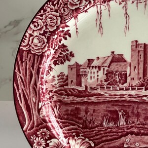 Op de afbeelding: Een decoratieve plaat met een bordeauxrode en witte afbeelding. De plaat heeft een gedetailleerde illustratie van een kasteelsc&egrave;ne met bloemenranden. De rand van de plaat heeft een zwarte rand.