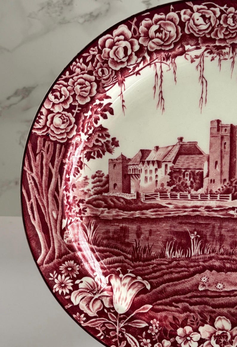 Op de afbeelding: Een decoratieve plaat met een bordeauxrode en witte afbeelding. De plaat heeft een gedetailleerde illustratie van een kasteelsc&egrave;ne met bloemenranden. De rand van de plaat heeft een zwarte rand.