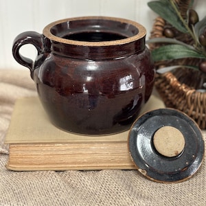 May include: A dark brown ceramic crock with a handle and a matching lid. The crock is round with a wide opening and sits on a vintage book. The lid is off to the side, revealing the interior.