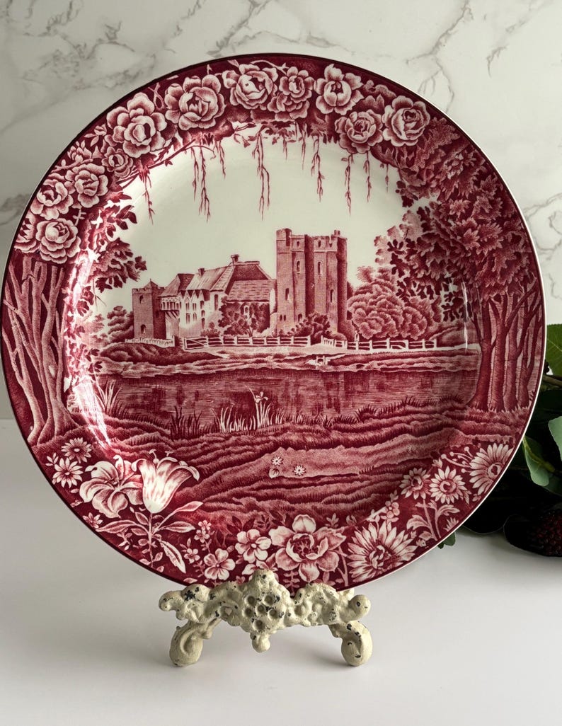 Op de afbeelding: Decoratieve bord met een bordeauxrood en wit ontwerp. Het bord heeft een gedetailleerde illustratie van een kasteelsc&egrave;ne, omgeven door bloemmotieven. De rand is versierd met rozen. Het bord wordt gepresenteerd op een witte decoratieve standaard.