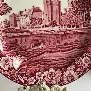 Op de afbeelding: Decoratieve plaat met een rood-witte kasteelsc&egrave;ne. De plaat heeft een gedetailleerde illustratie van een kasteel, water en een bloemenrand. De plaat wordt ondersteund door een witte, sierlijke standaard.
