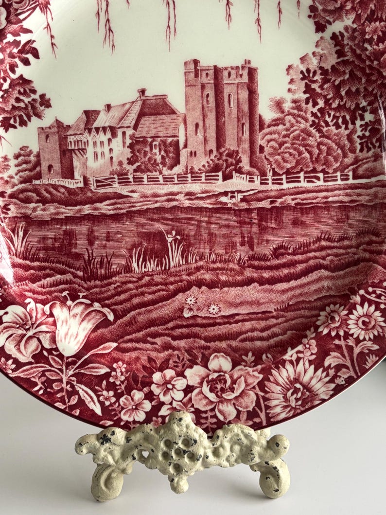 Op de afbeelding: Decoratieve plaat met een rood-witte kasteelsc&egrave;ne. De plaat heeft een gedetailleerde illustratie van een kasteel, water en een bloemenrand. De plaat wordt ondersteund door een witte, sierlijke standaard.