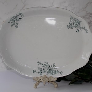 May include: Oval white serving platter with a scalloped edge. The platter has delicate teal floral designs in three places. The platter is displayed on a decorative stand, with greenery in the background.