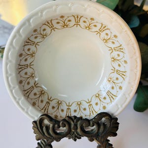 May include: A white ceramic bowl with a scalloped edge and gold floral pattern, displayed on a decorative bronze stand. The bowl's interior is plain, and the stand has an ornate, vintage design.