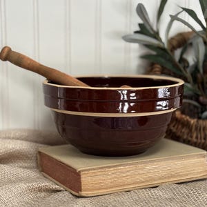 May include: A dark brown ceramic mixing bowl with a light brown rim and horizontal ridges. A wooden pestle rests inside the bowl. The bowl sits on a vintage book with aged pages, suggesting a rustic, farmhouse aesthetic.