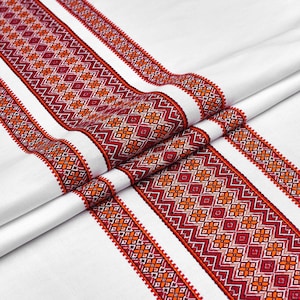 May include: White fabric with intricate embroidered patterns in red and orange. The design features geometric shapes and floral motifs, creating a detailed and textured appearance. The fabric is folded, showcasing the design's repeating pattern.