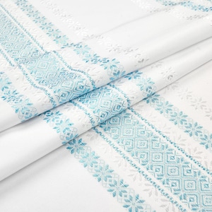 May include: White fabric with detailed turquoise and silver geometric and floral patterns. The design features stripes and borders with diamond and snowflake motifs. The fabric is folded, showing the texture and design.