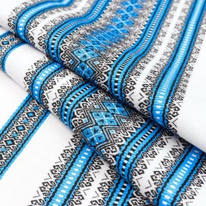 May include: Close-up of a folded white fabric with a detailed blue and black geometric pattern. The design features vertical stripes and diamond shapes, creating a textured appearance. The fabric is likely used for textiles or decorative purposes.