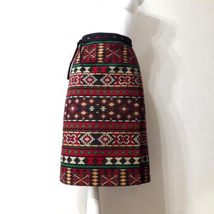 May include: A knee-length wrap skirt featuring a repeating geometric pattern in shades of red, black, green, and cream. The skirt has a solid black waistband with a side tie closure and a straight silhouette that falls just above the knee.