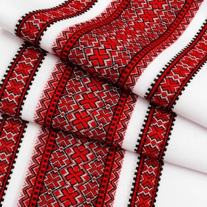 May include: White fabric with red and black embroidered geometric patterns. The design features repeating cross and zigzag motifs, creating a bold, traditional aesthetic. The fabric is folded, showing the intricate details of the embroidery.